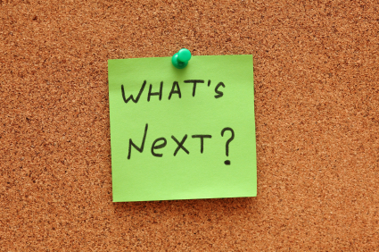 Green post-it note saying "What's next?" on corkboard (bulletin board). Close-up.